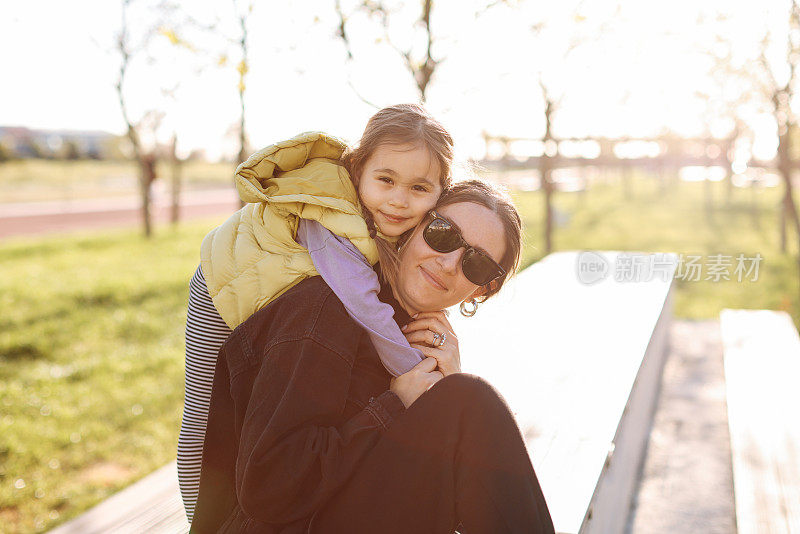 Young beautiful caucasian woman and cute daughter sitting on the bench in park in spring day